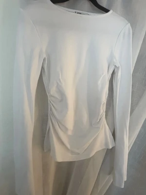 Rynkig långärmad tröja - Säljer en stilren vit långärmad topp med rynkade detaljer i sidan som ger en snygg form. Toppen har en rund halsringning och är tillverkad i ett mjukt material som känns skönt mot huden. Perfekt att matcha med nästan allt, i midjan har de slitsar. Helt oanvänd