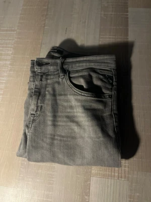 Grå slim fit jeans från replay - Snygga grå slim fit jeans från replay med klassisk femficksdesign och diskreta slitningar. Jeansen har dragkedjegylf och svarta detaljer vid fickorna. Perfekta för en avslappnad och trendig look. Stolek är 14åe= 166 cm! Modellen är Thad