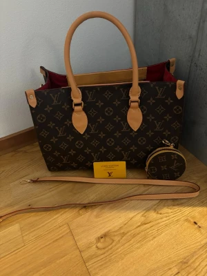 Louis Vuitton Monogram handväska - Säljer en klassisk Louis Vuitton handväska i brunt monogrammönster med beige läderdetaljer och rödfärgat foder. Väskan har två handtag, avtagbar axelrem och matchande rund liten väska. Guldiga metalldetaljer och dragkedja med Louis Vuitton-logga. Hyfsat sliten inuti väskan men utanpå som ny