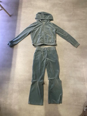 Mjukisset i turkost - Mysigt loungewear-set i grå velour med matchande hoodie och byxor. Hoodien har dragkedja, fickor och broderad detalj på ena fickan. Byxorna har dragsko i midjan och raka ben. Perfekt för chill dagar hemma.