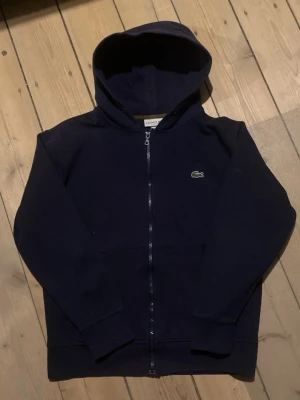 Mörkblå hoodie från Lacoste - Mörkblå hoodie från Lacoste med dragkedja framtill och klassisk krokodillogga på bröstet. Tröjan har huva och ribbade muddar vid ärmslut och nederkant. Perfekt för en avslappnad och stilren look.
