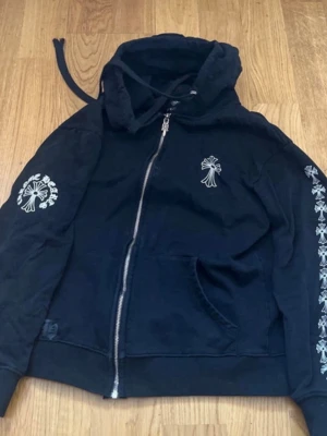 Svart hoodie från Chrome Hearts - Svart hoodie med dragkedja från Chrome Hearts. Har vita tryck med logga och kors på bröstet, ryggen och ena ärmen. Utrustad med huva och känguruficka. Snygg streetwear-stil med detaljerade tryck längs ärmen. kom dm för frågor 