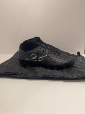 Svarta fotbollsskor från Skechers - Snygga svarta fotbollsskor från Skechers med hög krage och snörning. Skorna har en modern design med diskret mönster på ovandelen och Skechers-logga i silver på sidan. Tillverkade i syntetmaterial för bra grepp och kontroll på planen.
