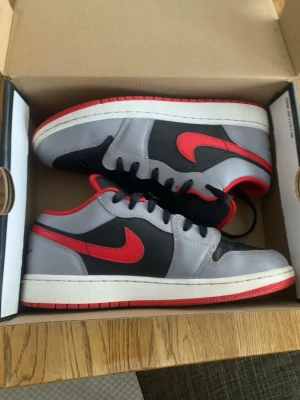 Nike Air Jordan 1 Low grå/röd - Snygga Nike Air Jordan 1 Low sneakers i grått och svart med röda detaljer och swoosh. Skorna har svart snörning, perforerad tåbox och röd yttersula. Insidan är fodrad i rött och materialet är en mix av läder och syntet. Perfekta för dig som gillar streetwear och klassisk sneakerstil.