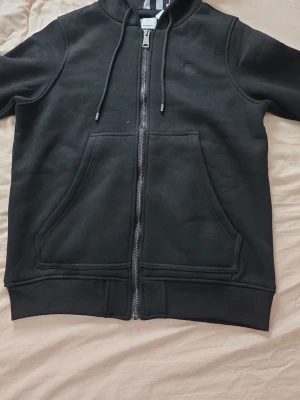 Svart zip-hoodie från Burberry - Svart hoodie med dragkedja från Burberry. Klassisk modell med huva och dragsko, samt två stora fickor framtill. Diskret Burberry-logga på bröstet och randigt innerfoder i huvan. Perfekt för en stilren och avslappnad look.