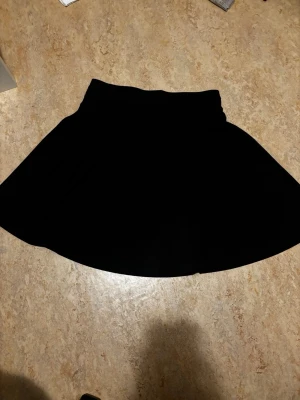 Svart kortkjol från Isolde - Säljer en svart kortkjol från Isolde. Kjolen har en enkel och klassisk design med lätt utställd form och bred linning. Perfekt att matcha med olika toppar för en snygg look.