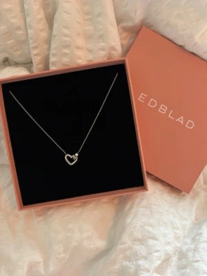 Halsband med hjärthänge från Edblad - Snyggt silverfärgat halsband från Edblad med ett hjärthänge dekorerat med små pärlor. Kedjan är tunn och elegant, perfekt för att lyfta vilken outfit som helst. Kommer i original ask.
