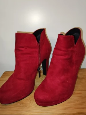 Röda stövletter från Tamaris - Snygga röda stövletter från Tamaris i mockaimitation med dragkedja på sidan och hög klack. Skorna har en stilren design och är fodrade med svart tyg invändigt. Perfekta för dig som vill sticka ut med färg och stil.
