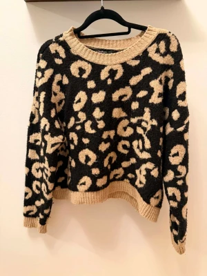 Leopardmönstrad stickad tröja - Cool stickad tröja med leopardmönster i svart och beige. Tröjan har rund halsringning och ribbstickade muddar vid ärmslut och nederkant. Perfekt för dig som vill sticka ut med ett trendigt djurmönster.