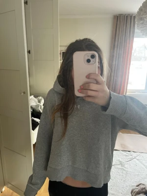 Grå hoodie från Under Armour - Snygg grå hoodie från Under Armour med croppad passform och klassisk huva. Hoodien har en liten liten målarfläck som visas på bild två men syns knappt.