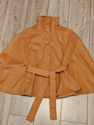 Camelbrun cape med bälte och hög krage - Stilren camelbrun cape med hög krage och knäppning framtill. Kappan har ett matchande bälte i midjan för snygg siluett och är tillverkad i ett mjukt tyg som påminner om ull. Perfekt för dig som vill ha en trendig och tidlös look. Poncho, färgen ser lite annorlunda ut på bilden. Strl M-S, helt ny aldrig använd