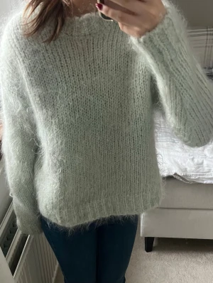 Ljusgrön fluffig stickad tröja - Mysig ljusgrön stickad tröja med fluffig känsla och grovstickat mönster. Tröjan har rund halsringning och långa ärmar, perfekt för kyliga dagar. Materialet ser ut att vara en mjuk blandning med mohair eller liknande.