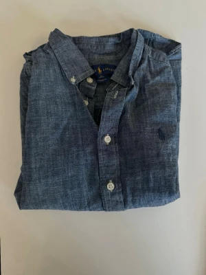 Blå skjorta från Ralph Lauren - Klassisk blå skjorta från Ralph Lauren med button-down krage och broderad logga på bröstet. Skjortan har långa ärmar, vita knappar och är tillverkad i mjuk bomull. Perfekt för en avslappnad men ändå stilren look.