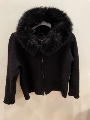 ALESSA cardigan/Svart teddyjacka med fuskpäls - Säljer en svart ALESSA teddyjacka med fluffig fuskpäls runt huvan. Jackan har dragkedja framtill, ribbade muddar mycket mysig,. Perfekt för kalla dagar när du vill vara både varm och snygg.
