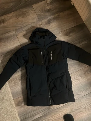 Mörkblå svart, vita detlajer vindjacka från Sail Racing - Snygg mörkblå vindjacka från Sail Racing med huva och dragkedja framtill. Jackan har två sidofickor och en bröstficka med dragkedja samt logga på bröstet. Tillverkad i slitstarkt material som står emot vind och väta. Perfekt för blåsiga dagar.