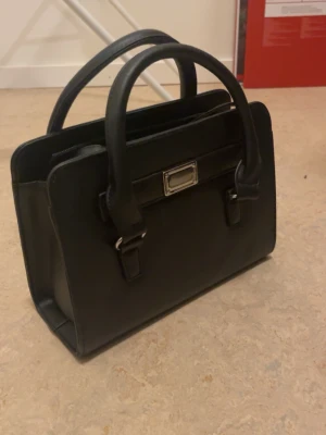 Svart stilren handväska i läderlook - Snygg svart handväska med rektangulär form och dubbla handtag. Väskan har silverfärgade metalldetaljer och en dekorativ spänne framtill. Perfekt för dig som gillar en clean och klassisk stil. Materialet ser ut som konstläder. Med följer ett långt band med 