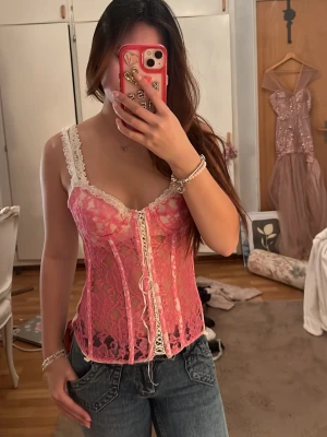 Rosa spetskorsett med vita detaljer - Säljer en snygg rosa korsett i genomskinlig spets med vita spetsdetaljer och smala axelband. Framsidan har dekorativa snörningar och kupor med lätt vaddering. Perfekt för att lyfta en outfit med en feminin touch.