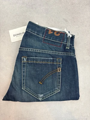 Blå jeans från Dondup - Snygga blå jeans från Dondup med klassisk femficksdesign och raka ben. Jeansen har kontrastsömmar och en unik detalj på bakfickan. Tillverkade i slitstarkt denim med en tvättad finish och läderpatch bak i midjan.