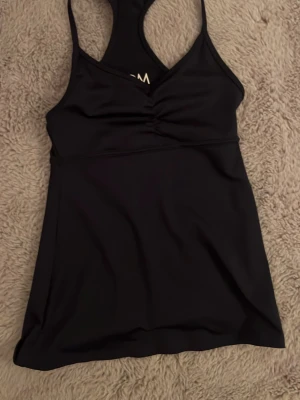 Svart sportlinne från DOM - Snyggt svart sportlinne från Drop of Mindfulness med brottarrygg. Tillverkat i stretchigt material av 86% polyester och 14% elastan. Linnet har en lätt rynkning framtill och sitter tajt för en sportig look.