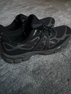 Svarta Asics sneakers med grå detaljer - Säljer ett par svarta Asics sneakers med grå detaljer och reflekterande partier. Skorna har en chunky sula, snörning med mönstrade skosnören och Asics-logga i rött på plösen. Materialet är en mix av mesh och syntet för en sportig look. Pris kan diskuteras 