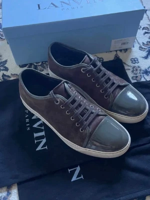 Bruna sneakers från Lanvin i mocka - Snygga bruna sneakers från Lanvin med ovandel i mocka och tåparti i blankt grått material. Klassisk låg modell med vita sulor och matchande bruna skosnören. Perfekta för dig som vill ha en stilren och exklusiv sneaker.