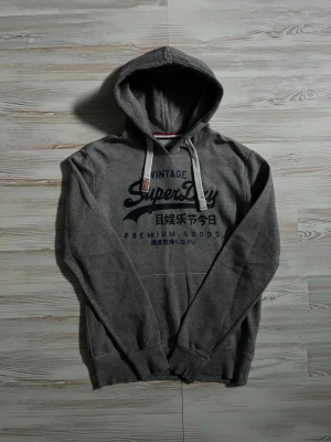 Grå Superdry hoodie med tryck - Snygg grå hoodie från Superdry med stort blått tryck på bröstet och japanska tecken🤩 Liten defekt på bröstet men det är inte nåt man tänker på😅Perfekt för en avslappnad streetstyle-look. Hör av er vid funderingar🙌🏻