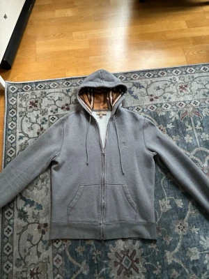 Grå burberry hoodie - Snygg grå hoodie med dragkedja från Burberry. Klassisk huva med justerbara snören och ikoniskt rutigt foder på insidan. Två stora fickor framtill och diskret Burberry-logga på bröstet. Perfekt för en chill och stilren look.