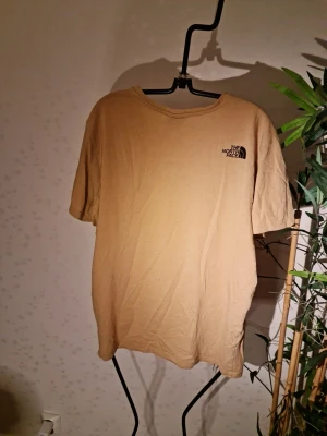 Beige t-shirt från The North Face - Snygg beige t-shirt från The North Face med klassisk logga tryckt på bröstet. T-shirten har rund halsringning och korta ärmar. Perfekt basplagg i mjuk bomull som funkar till det mesta.