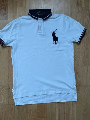 Vit pikétröja från Polo Ralph Lauren - Vit pikétröja från Polo Ralph Lauren med klassisk krage i marinblått med röda och vita ränder. Stor broderad logga på bröstet och knäppning framtill. Tröjan har korta ärmar med matchande kantband och är i bomull.