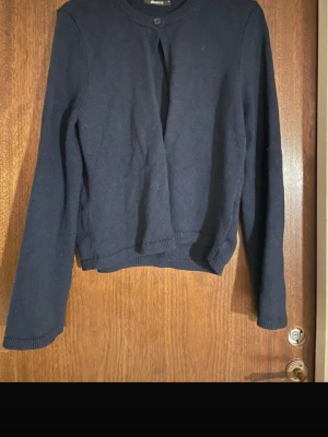 Mörkblå kofta  - Mörkblå kofta med rund halsringning och knappstängning upptill. Långärmad modell i mjukt stickat material med ribbade muddar vid ärmslut och nederkant. Enkel och stilren design som passar till det mesta.