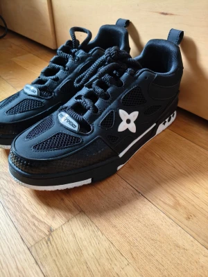 Svarta Louis Vuitton sneakers - Svarta sneakers från Louis Vuitton med vit blomdetalj på sidan och vita inslag på sulan. Skorna har meshpaneler, svarta skosnören och robust sula. Snygg och modern design med tydliga streetwear-vibbar. Perfekta för dig som gillar exklusiva märken och unik stil.
