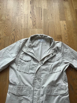 Beige vindjacka från John Henric - Stilren beige vindjacka från John Henric med fyra praktiska fickor framtill och hög krage med tryckknappar. Jackan har dragkedja och är tillverkad i ett lätt material som passar perfekt för blåsiga dagar. Enkel och clean design som funkar till det mesta. Bara använd ett fåtal gånger.