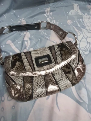 Vintage väska i ormmönster - Från River Island. Guess liknande handväska i metallic silvrigt ormmönstrat material med bred axelrem och stora metallringar. Väskan har ett rektangulärt spänne framtill och detaljer i svart. Perfekt för dig som gillar statement-accessoarer och unika detaljer.