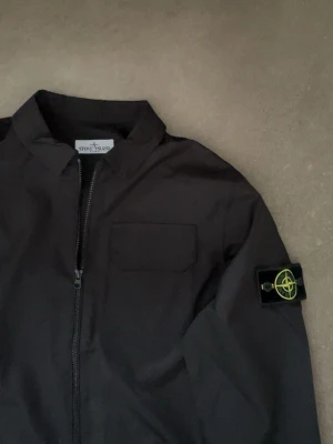 Svart overshirt från Stone Island - Stilren svart overshirt med dragkedja från Stone Island. Klassisk patch på vänster ärm och bröstficka framtill. Perfekt för lager-på-lager och enkel att matcha med andra plagg. Overshirt är M men passar även S. Pris kan diskuteras vid snabb affär