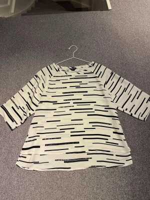 Vit blus med svart mönster från Lindex - Snygg vit blus från Lindex med svart grafiskt mönster i form av linjer. Blusen har trekvartsärm och en avslappnad passform. Perfekt till jeans eller kjol för en trendig look.