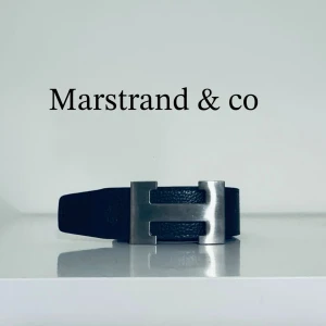 Svart skärp från Marstrand & co - Snyggt svart skärp från Marstrand & co med en stor, silverfärgad metallspänne i form av ett H. Bältet är gjort i skinn och har en stilren och klassisk look som passar till många outfits.