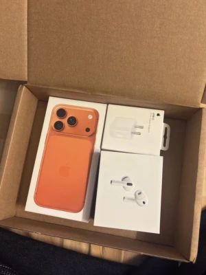 Iphone 17 pro max 512GB - Säljer en orange iPhone från Apple med snygg design och rundade kanter. Telefonen är tillverkad i glas och aluminium. Medföljer gör även en laddare och ett par vita trådlösa hörlurar (AirPods). Perfekt för dig som gillar stilrena och moderna prylar.