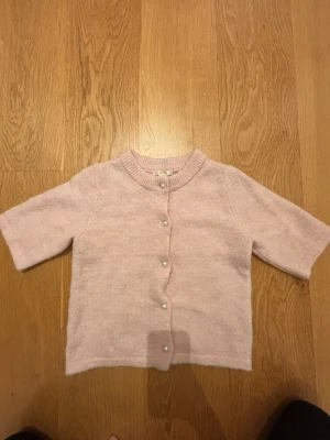 Ljusrosa kofta från Gina Tricot Young - Supersöt ljusrosa kofta från Gina Tricot Young i storlek 146/152 med korta ärmar och pärlknappar framtill. Koftan är stickad i mjukt material och har ribbad halsringning. 