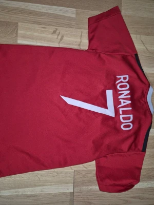  Barn Manchester United Ronaldo tröja   - Röd Manchester United matchtröja från Adidas med tryck 'Ronaldo 7' på ryggen och klubbmärke på bröstet. Tröjan har vita och svarta detaljer, korta ärmar och TeamViewer som sponsortryck. Perfekt för fotbollsfans som vill visa sitt stöd. Ej original 