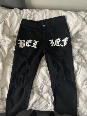 Svarta ripped jeans med text - Säljer ett par svarta jeans med stora vita tryck i gotisk stil där det står 'BELIEF' över låren. Jeansen har flera slitningar och hål på framsidan för en edgy look. Perfekta för dig som gillar streetwear och vill sticka ut.