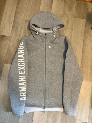 Grå hoodie från Armani Exchange - Snygg grå hoodie från Armani Exchange med dragkedja och stor vit logga längs ena ärmen. Den är strl M men passar även S. Nypris 5000kr. Om några frågor eller prisförslag så är det bara o hojta till 