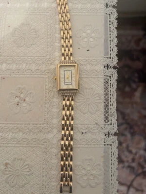 Guldfärgad elegant klocka Omax - Stilren guldfärgad klocka från Omax med rektangulärt ur och länkarmband i metall. Urtavlan är vit med romerska siffror och detaljer i guld. Klassisk och elegant design som passar till många stilar.