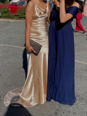 Champagnefärgad långklänning med slits - Elegant långklänning i champagnefärgad satin med smala axelband och draperad urringning. Klänningen har en hög slits framtill och ett släp baktill för extra effekt. Perfekt för bal eller festliga tillfällen. Storlek 36. Köpt för 1500kr från zalando. Haft på mig den en gång 