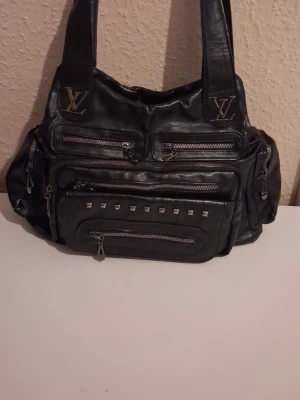 Svart Louis Vuitton handväska i skinn  - Cool svart handväska från Louis Vuitton i skinn med flera dragkedjefack och nitar framtill. Väskan har breda axelband och LV-loggor i metall. Perfekt för dig som gillar en edgy och praktisk look med mycket plats för dina grejer.