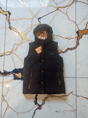 Helt Ny Svart dunväst med huva Burberry - Snygg svart dunväst från Burberry med avtagbar huva och klassiskt rutigt foder. Västen har dragkedjor på fickorna och tryckknappar framtill. Perfekt för lager på lager och streetstyle. Märkespatch på bröstet och exklusiv känsla.