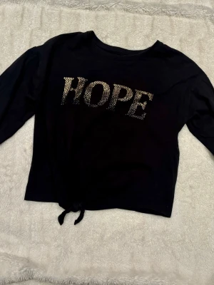 Svart croppad topp med HOPE-tryck - Svart långärmad croppad topp med knyt i midjan och texten HOPE framtill i glittriga detaljer. Perfekt för en trendig och avslappnad stil. Perfekt skick, använd endast ett par gånger.