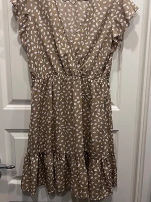 Beige omlottklänning från SHEIN - Snygg beige kortklänning från SHEIN med vitt prickigt mönster. Klänningen har omlottmodell, volangdetaljer vid ärmar och nederkant samt resår i midjan för en bekväm passform. Perfekt för sommarens alla tillfällen!