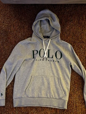 Grå hoodie från Polo Ralph Lauren - Snygg grå hoodie med gröna detaljer från Polo Ralph Lauren. Huva med vita snören och liten broderad logga på ärmen. Tillverkad i mjuk bomullsblandning som känns skön mot huden. Perfekt för en avslappnad och trendig look.