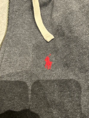 Grå hoodie från Polo Ralph Lauren - Mörkgrå hoodie med dragkedja från Polo Ralph Lauren. Klassisk röd logga broderad på bröstet, vita snören i huvan och två fickor framtill. Tillverkad i mjuk bomullsblandning, perfekt för en avslappnad stil.