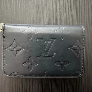 Svart plånbok  - Ny, liten svart plånbok med silvrig dragkedja och mönster på. Fack för kort mm.. Kommer med box också, om man vill ha. Ca 10.5 cm och 7 cm hög.